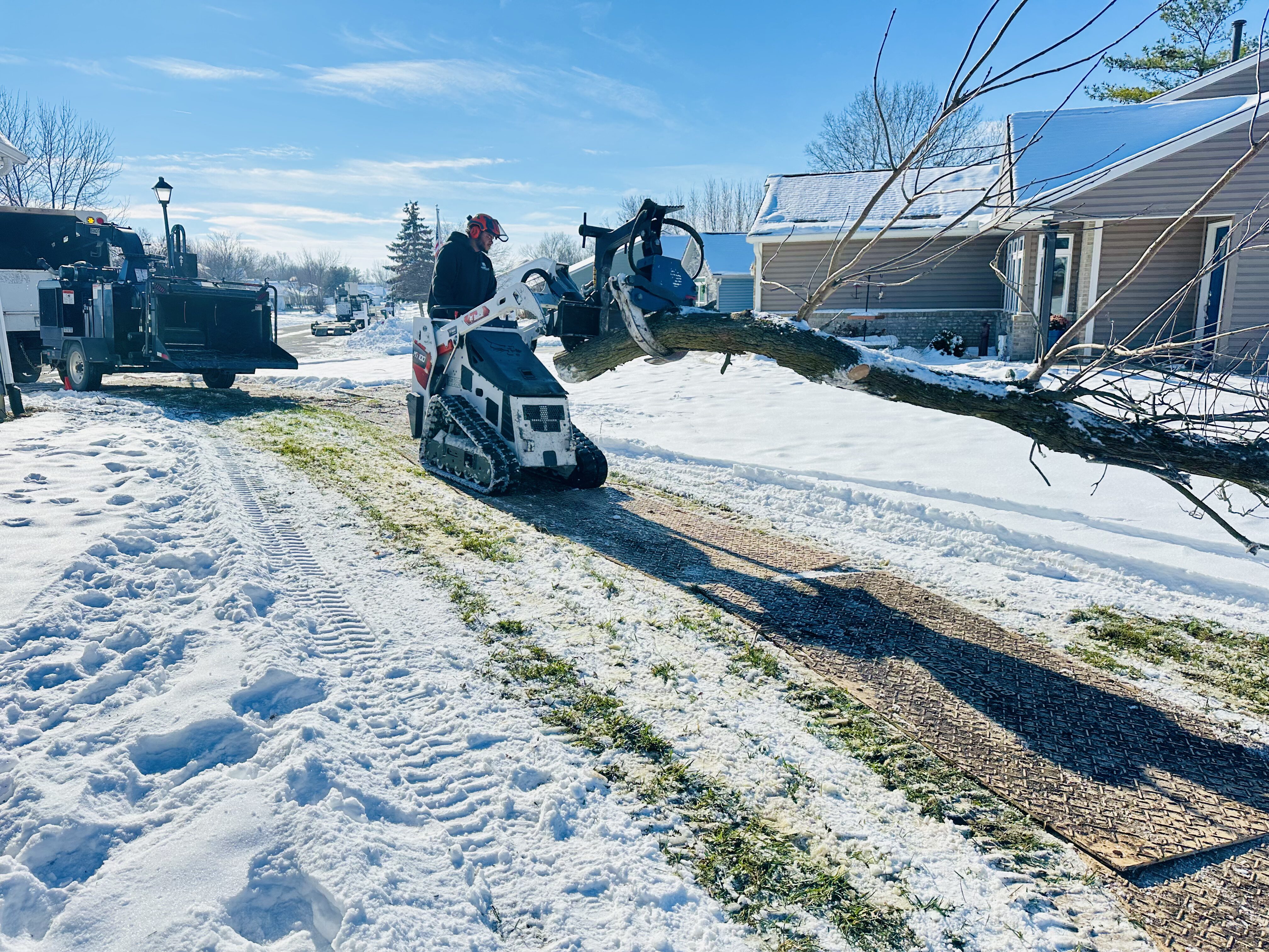 In a snow-covered residential area, a white compact track loader uses its grapple to lift a large tree limb off a driveway. The operator is wearing high-visibility gear to safely clear the winter storm debris