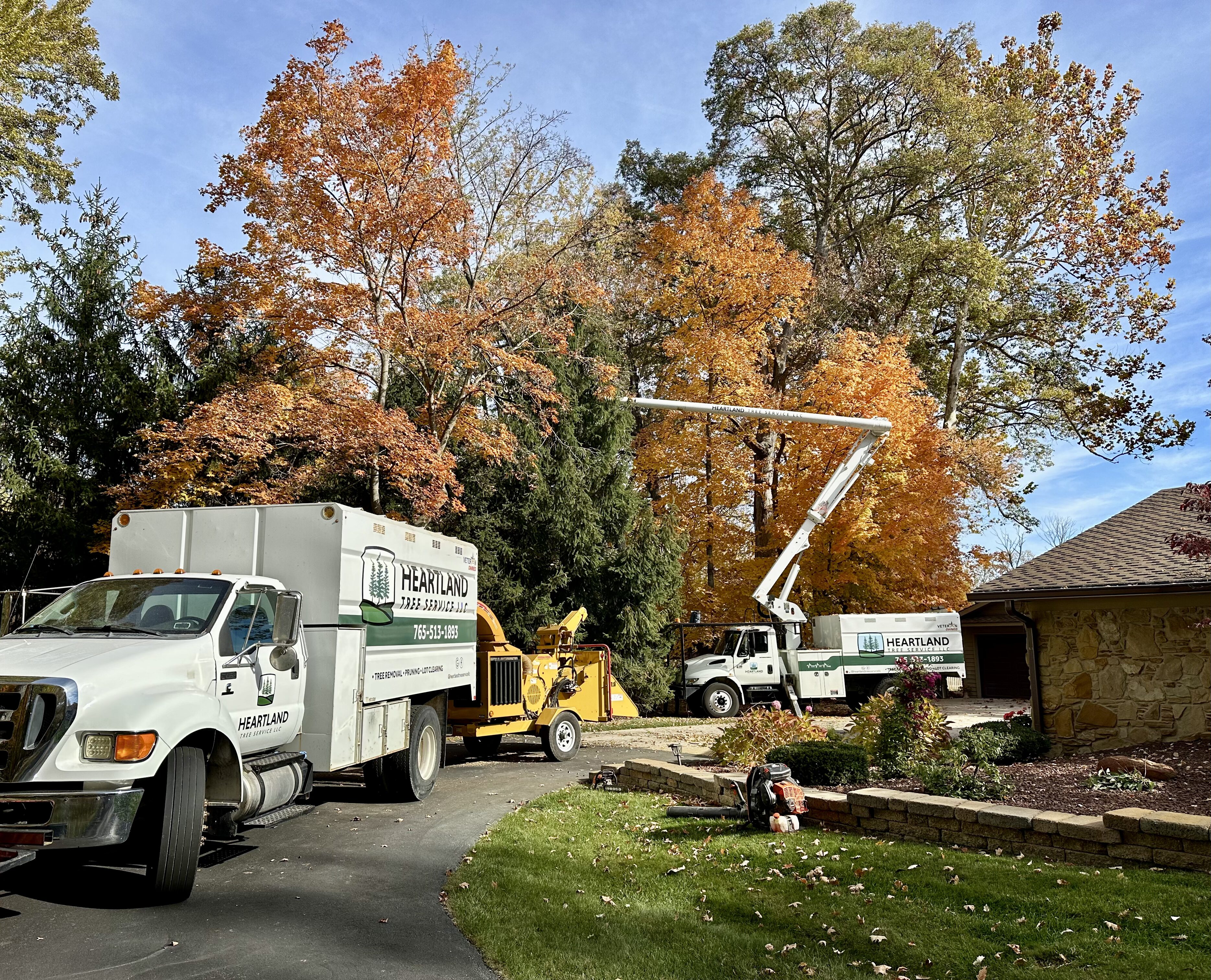 A white professional tree service truck and a wood chipper are parked in a residential driveway. In the background, a long mechanical lift arm extends high into a canopy of vibrant orange and yellow autumn trees