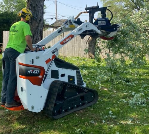 A professional arborist in a high-visibility lime green shirt and a yellow safety helmet operates a white Bobcat MT100 mini track loader. He is using a grapple attachment with a patriotic American flag decal to lift and clear fallen tree branches from a grassy residential yard