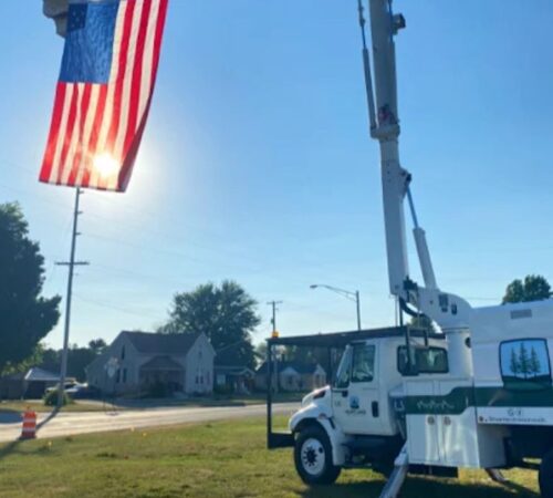 A Heartland Tree Service bucket truck is parked on a grassy verge with its mechanical arm raised high. In the foreground, a large American flag hangs suspended from the arm, backlit by the bright afternoon sun